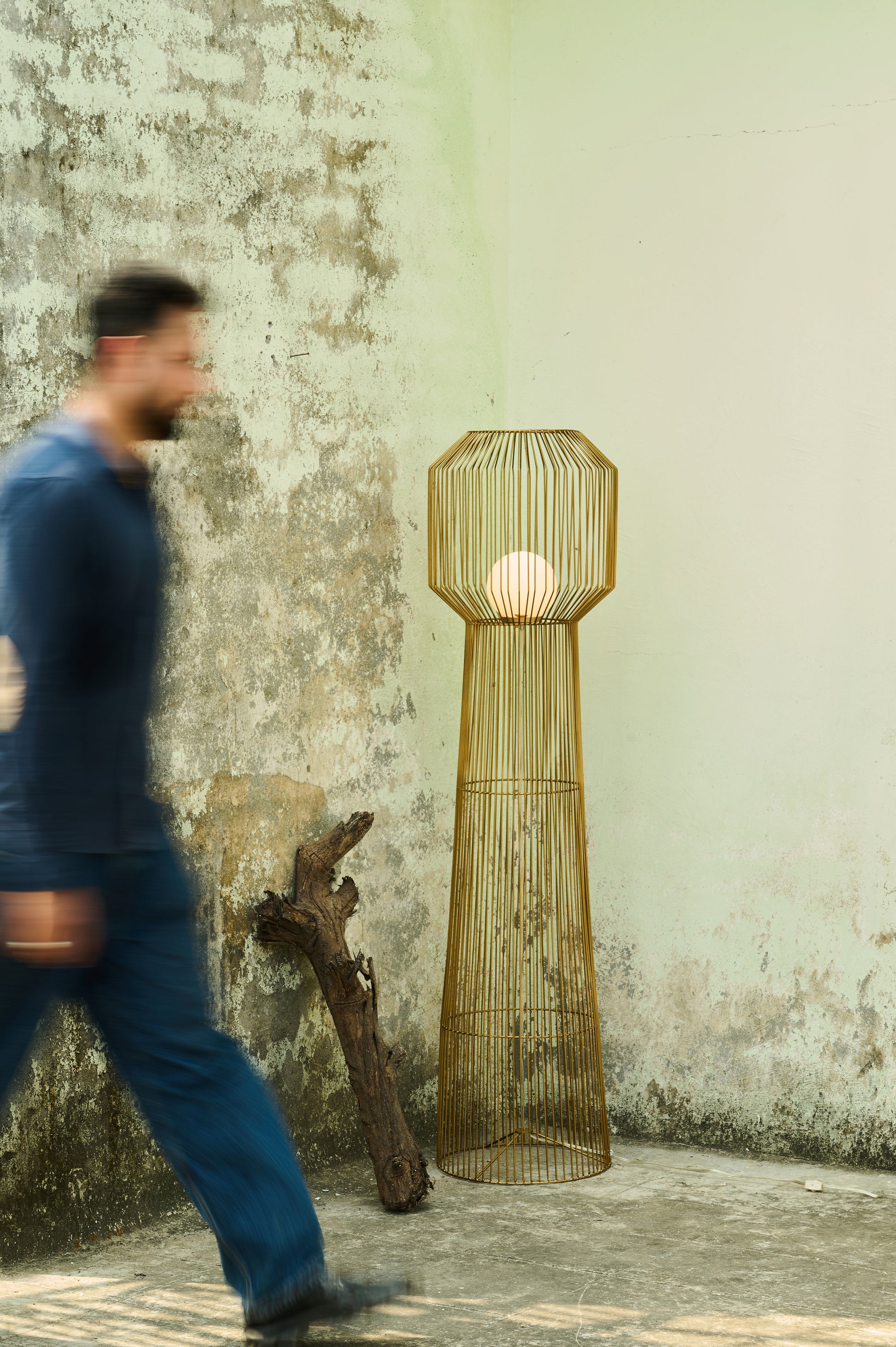 Gold wire floor lamp against a textured wall with a person walking by
