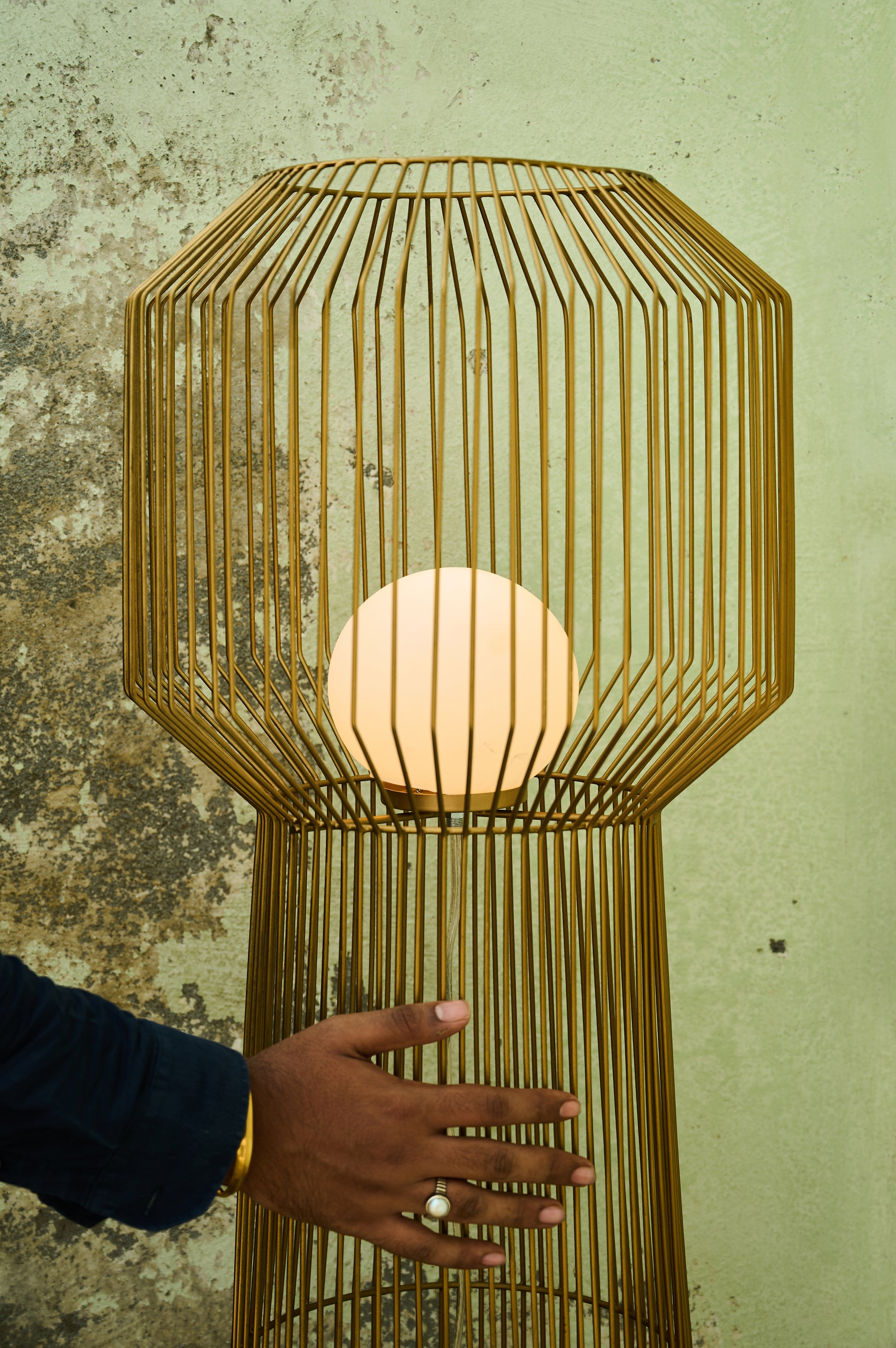 Gold cage-style lamp with a hand reaching towards it against a textured wall.