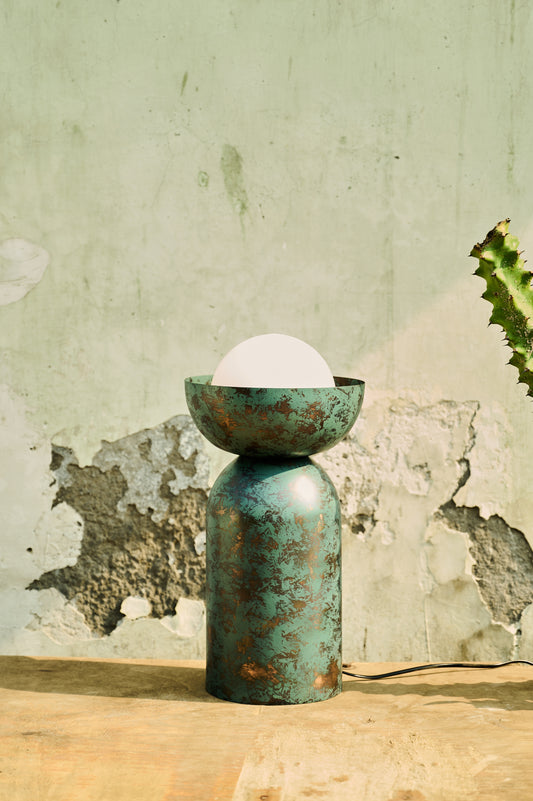 Green ceramic lamp with a white bulb on a wooden surface against a textured wall.