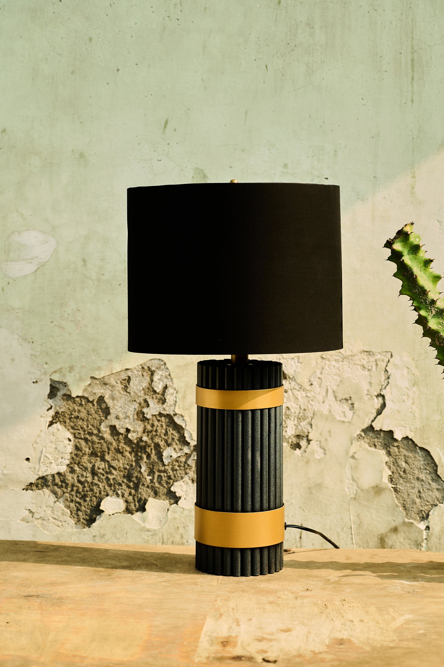 Table lamp with black shade and ribbed black and gold base on a wooden surface with a textured wall background.