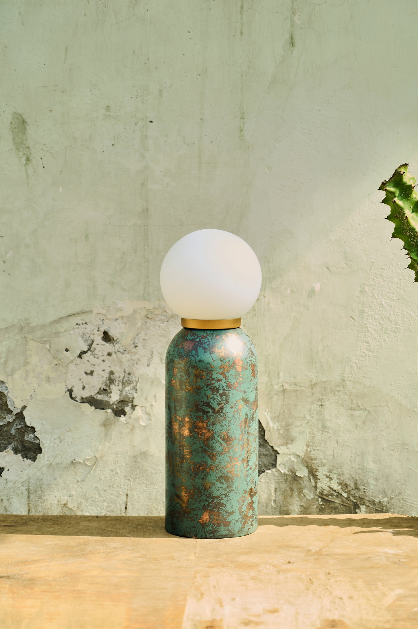 Table lamp with a textured green base and white globe shade on a wooden surface with a rustic wall background.