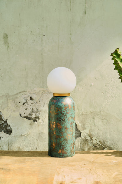 Table lamp with a textured green base and white globe shade on a wooden surface with a rustic wall background.