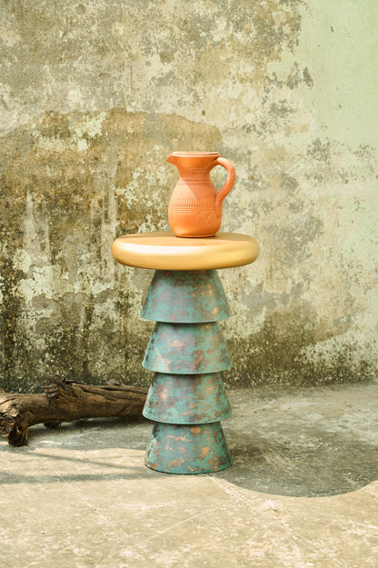 Terracotta pitcher on a stacked metal stool against a textured wall.