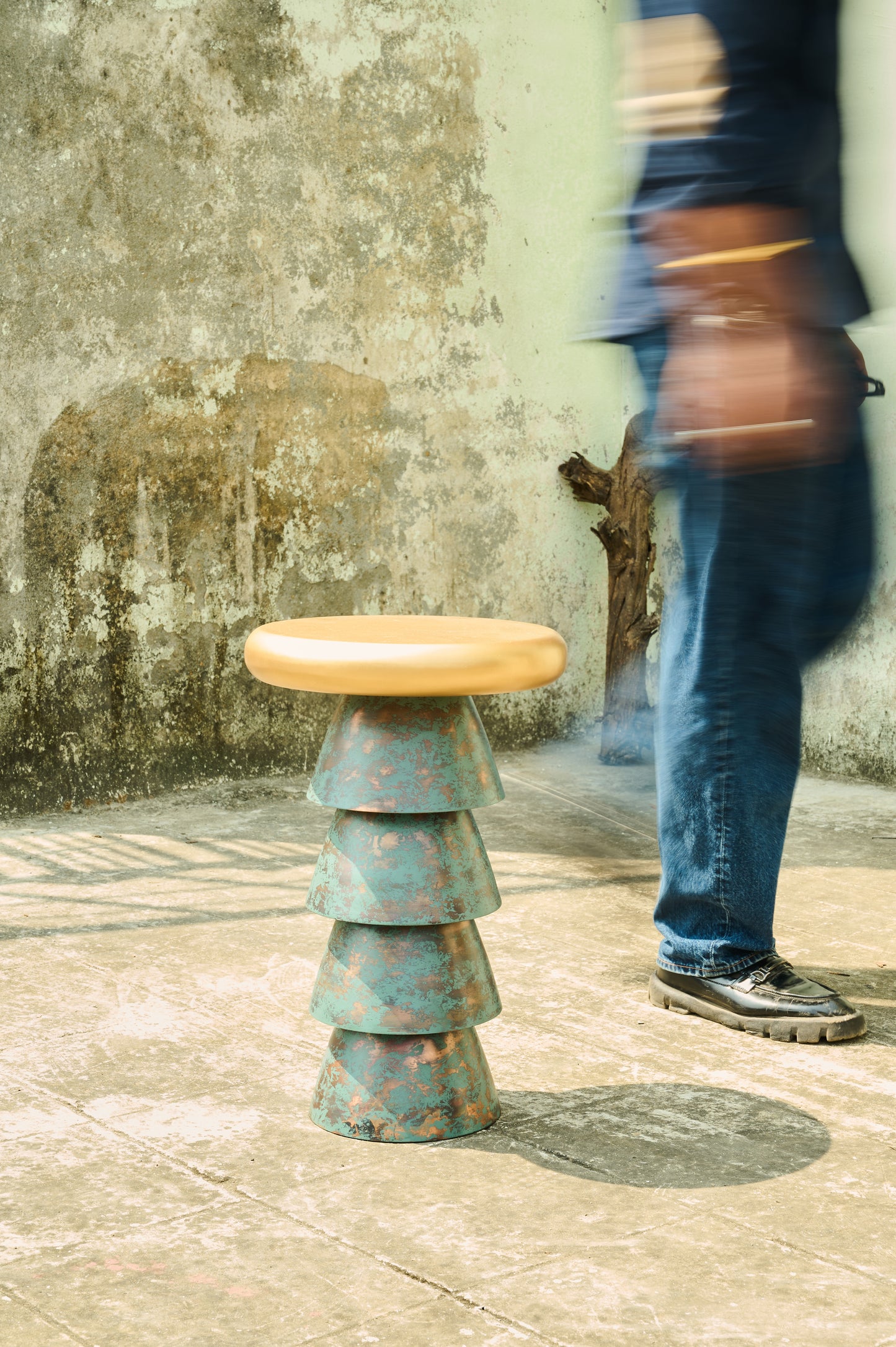 Stacked metal stool with a wooden top on a textured floor with a person walking by.