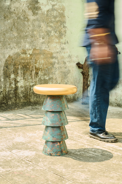 Stacked metal stool with a wooden top on a textured floor with a person walking by.