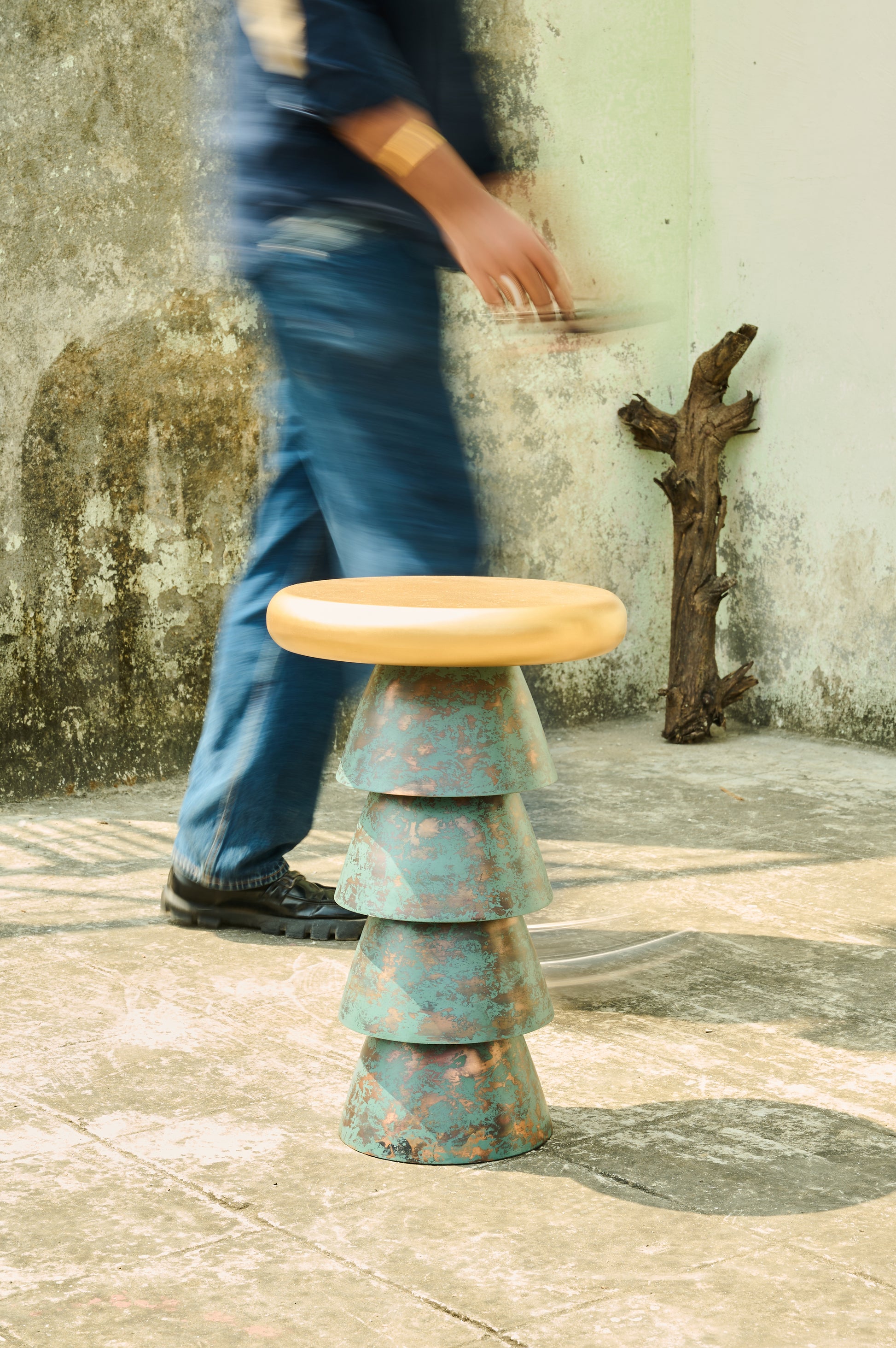 Person walking past a unique, tiered stool with a wooden top against a textured wall.