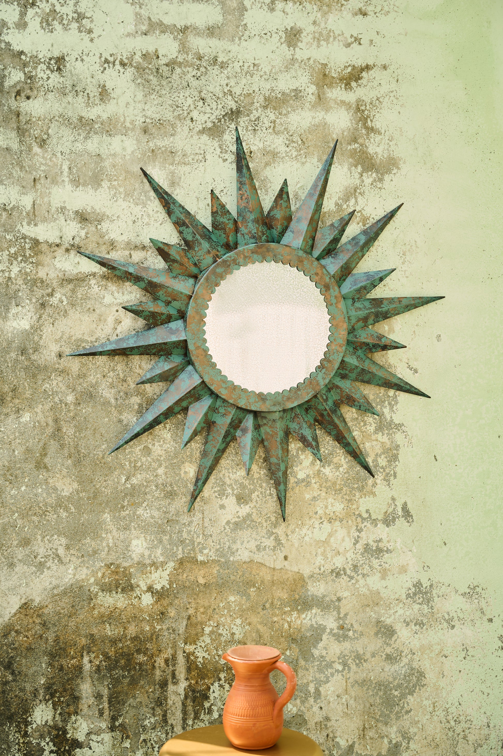Decorative sun-shaped mirror on a textured wall with a small orange pitcher below.
