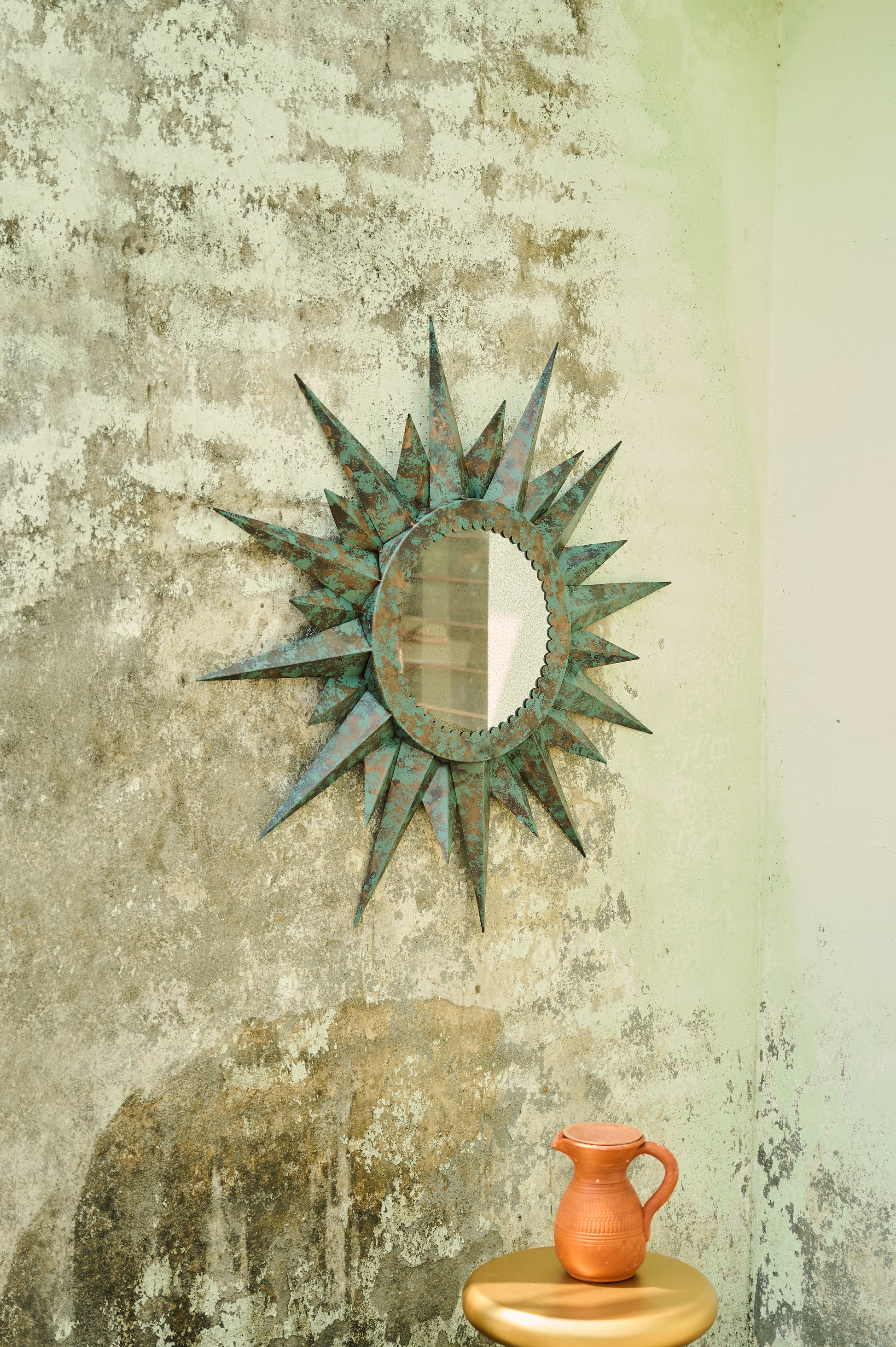 Decorative sunburst mirror on a textured wall with a small orange pitcher on a wooden stool.