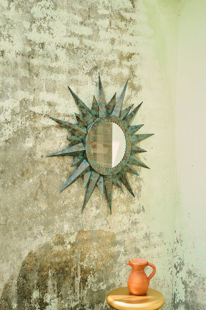 Decorative sunburst mirror on a textured wall with a small orange pitcher on a wooden stool.