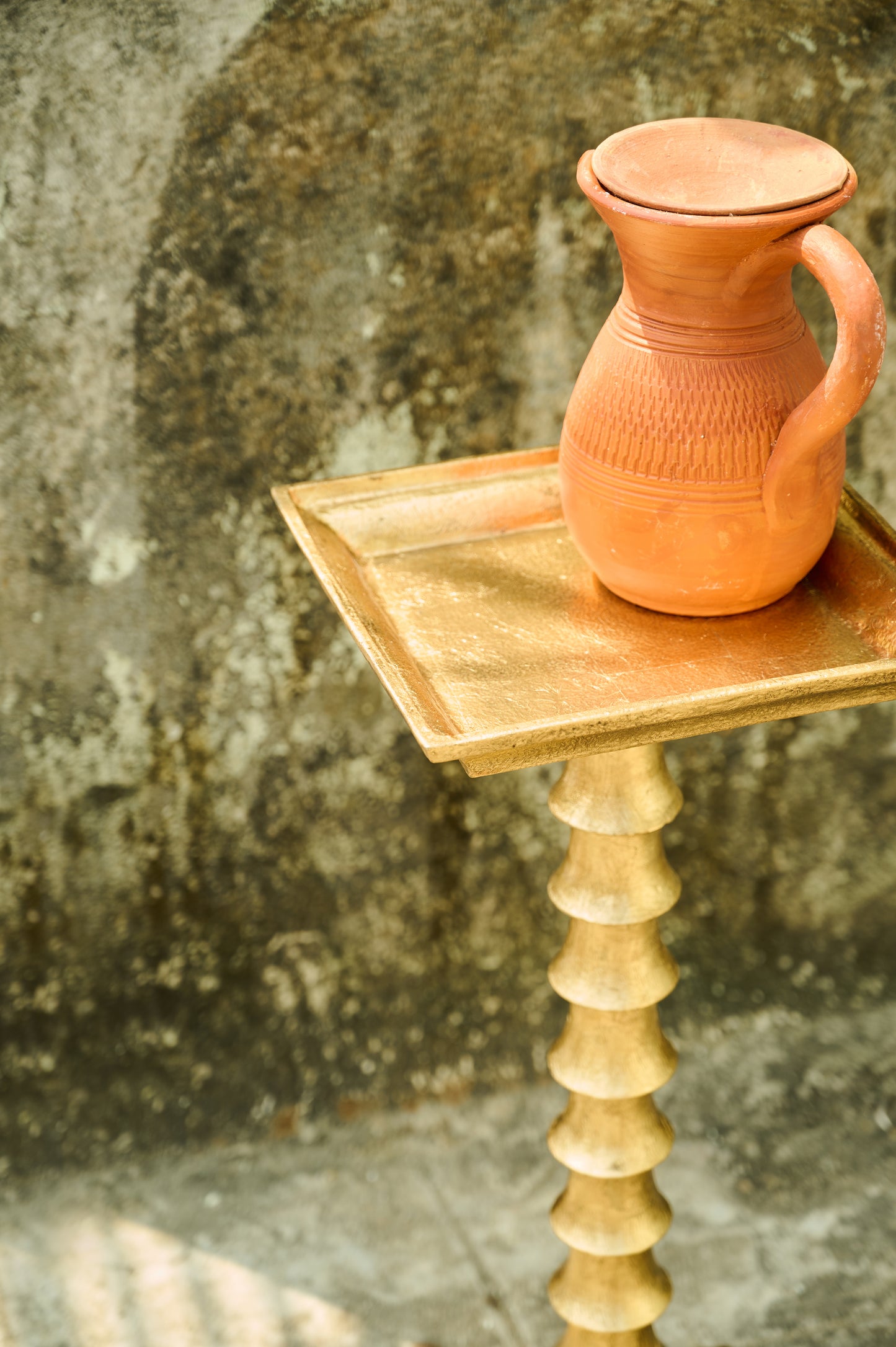 Terracotta pitcher on a wooden stand against a textured wall.