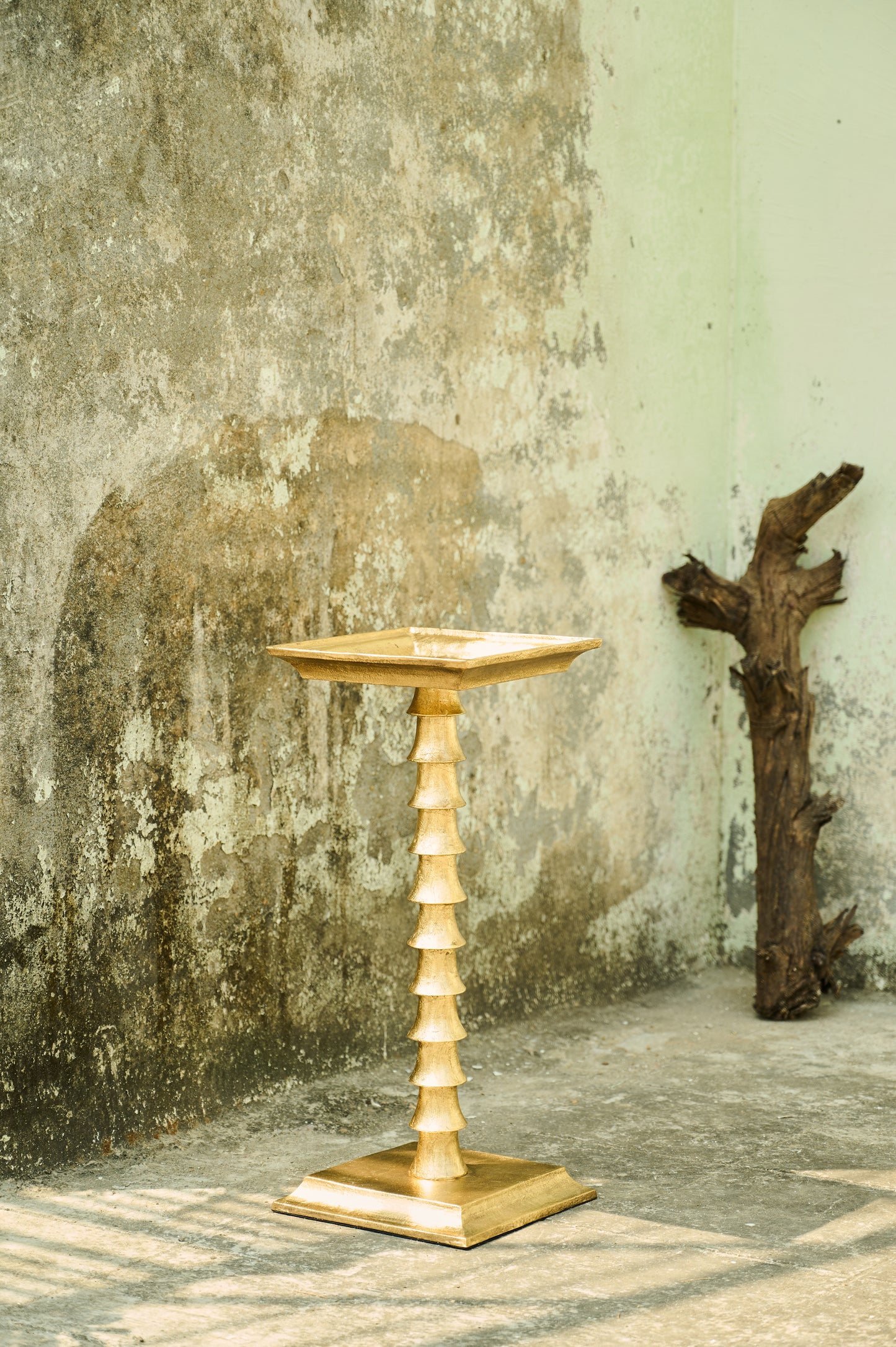 Gold pedestal table against a textured wall with a piece of driftwood.