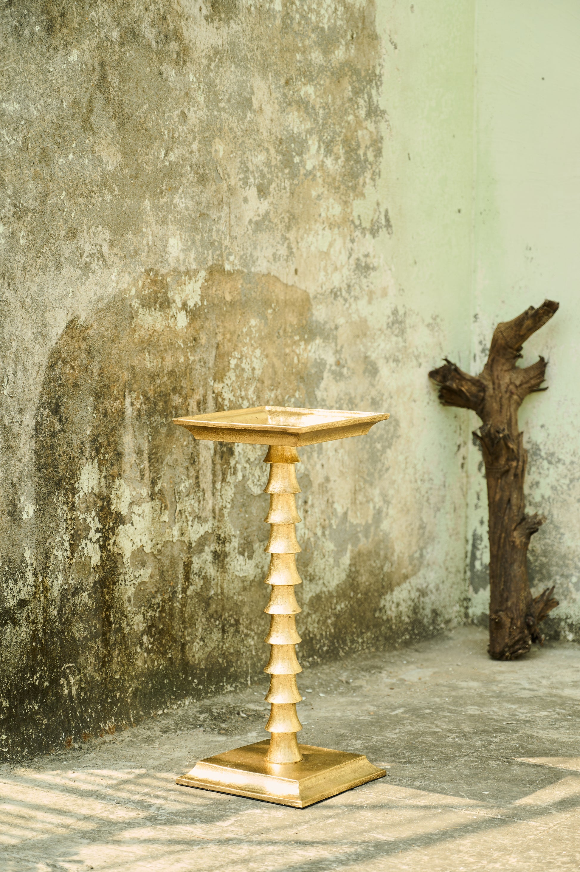 Gold pedestal table against a textured wall with a piece of driftwood.